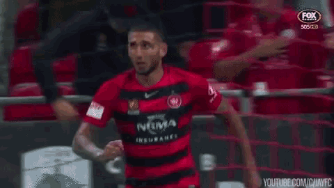 a man in a red and black soccer jersey is standing in front of a fence .
