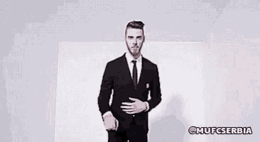 a man in a suit and tie is standing in front of a white wall and pointing at the camera .