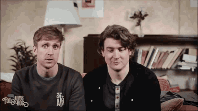 two men are sitting on a couch and one is wearing a shirt that says arms of the tulle hog