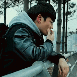 a young man in a leather jacket is leaning on a railing and smoking a cigarette .