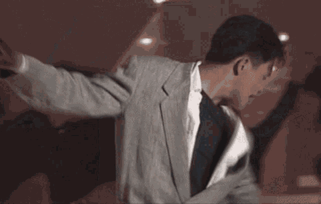 a man in a grey jacket and white shirt is waving his hand in the air