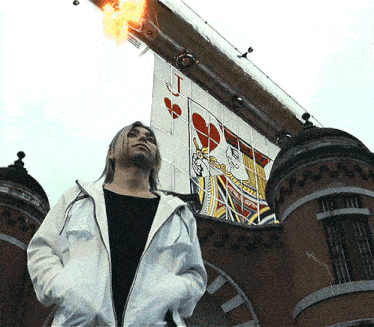 a woman in a white jacket stands in front of a giant jack of hearts playing card