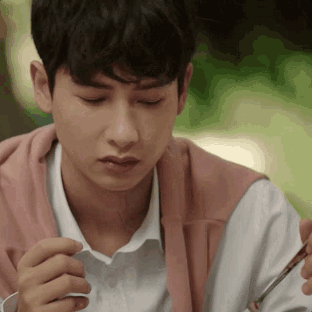 a young man in a white shirt and pink sweater is eating something with a fork and knife .
