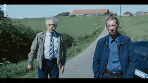 two men standing next to each other on a road with the letter g on the side of the car