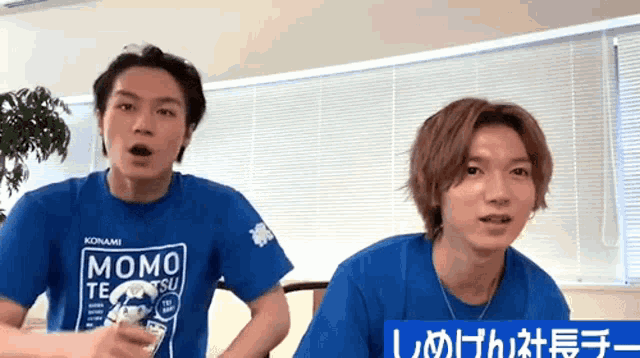 two young men wearing blue momo t-shirts are standing next to each other in a room .