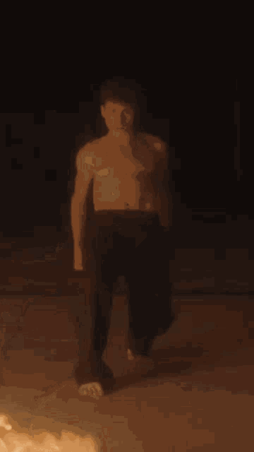 a man without a shirt is standing in front of a fire in the dark .