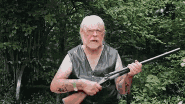 a man in a leather vest is holding a shotgun in his hands in the woods .