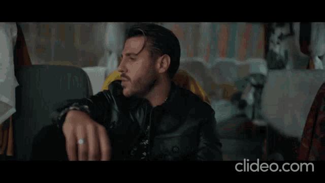 a man in a black leather jacket is sitting on a couch with his hand on his knee .
