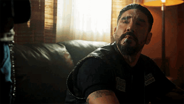 a man sitting on a couch smoking a cigarette and wearing a vest that says sons of anarchy on it