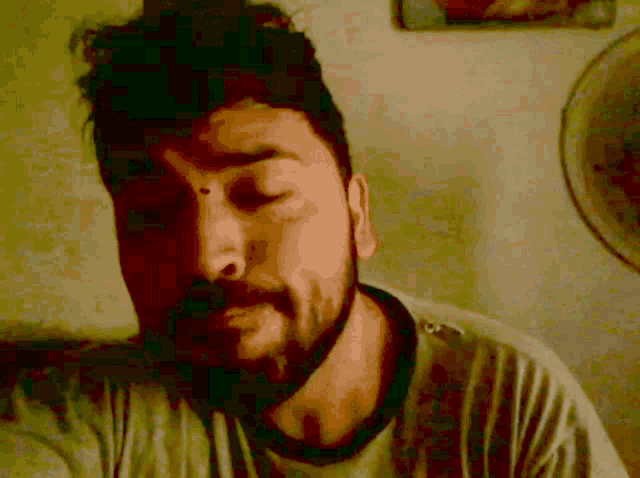 a man with a beard and a gray shirt looks at the camera with his eyes closed
