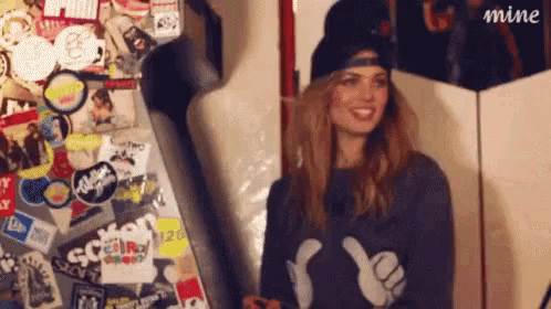 a woman giving a thumbs up in front of a wall covered in stickers and a sign that says mine