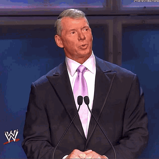 a man in a suit and tie stands at a podium with a w logo on the bottom
