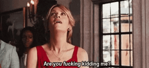 a woman in a red dress is standing in front of a window and asking are you fucking kidding me ?