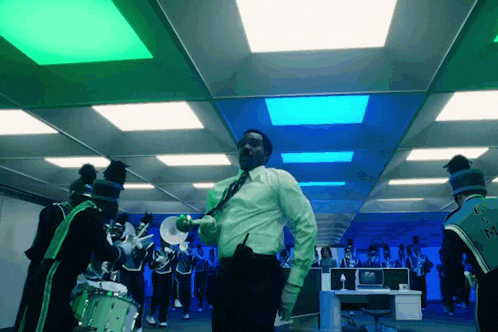 a man in a green shirt and tie stands in front of a marching band with the letter m on the back of their uniform