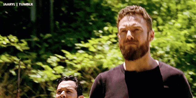 a man with a beard wearing a black shirt is standing in the woods .