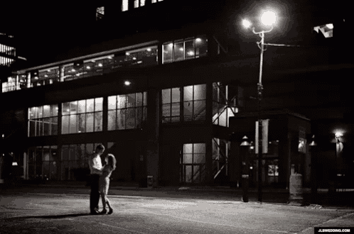 a black and white photo of a man and woman kissing in front of a large building