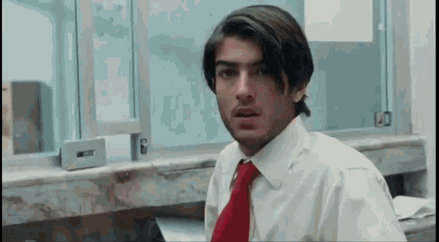 a man in a white shirt and red tie is sitting at a desk in front of a window .