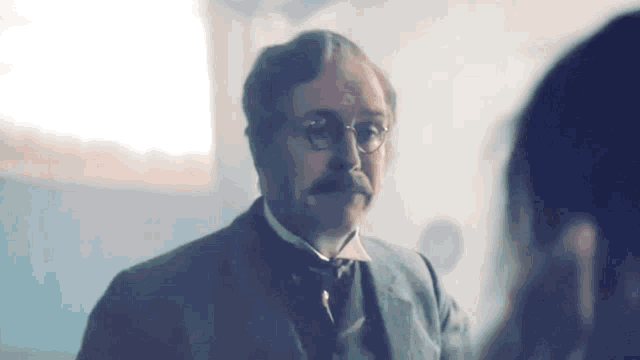 a man in a suit and tie with glasses and a mustache is talking to a woman .