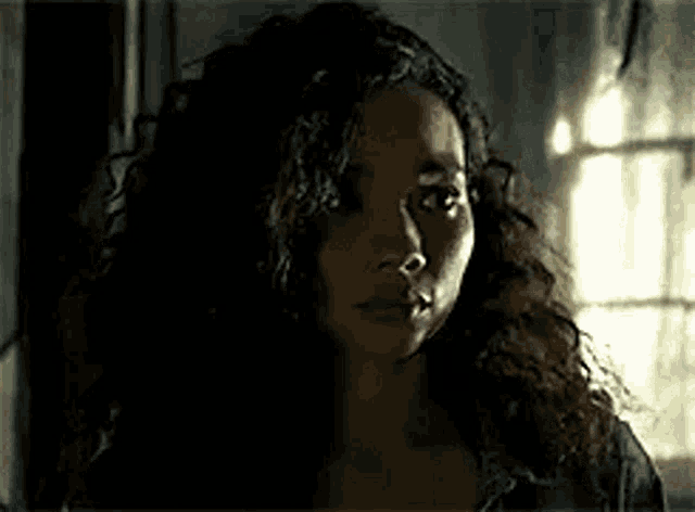 a woman with curly hair is standing in front of a window looking at the camera .