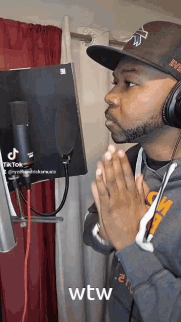 a man wearing headphones and a hat is standing in front of a microphone with his hands folded in prayer