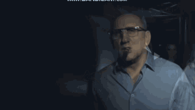 a man wearing glasses and a blue shirt is smoking a cigar in a dark room