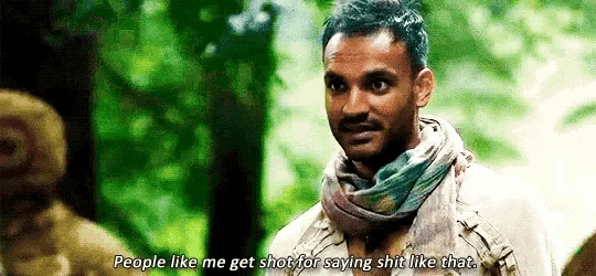a man with a scarf around his neck is talking to a group of people in a forest .