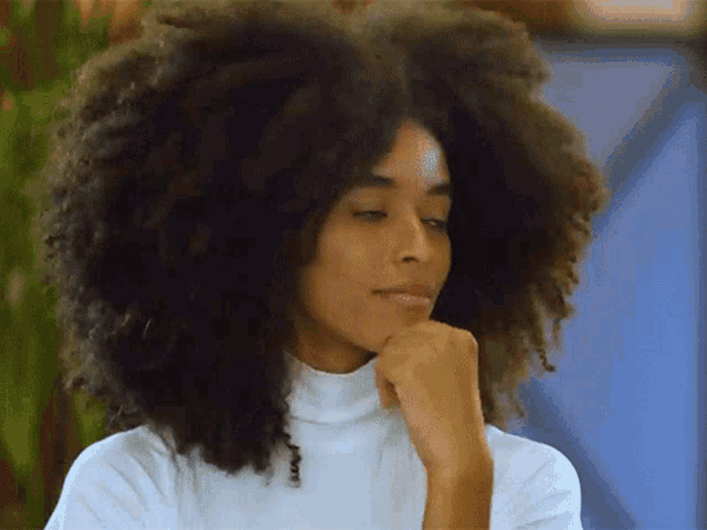 a woman with a very large afro is wearing a white turtleneck sweater and has her hand on her chin .