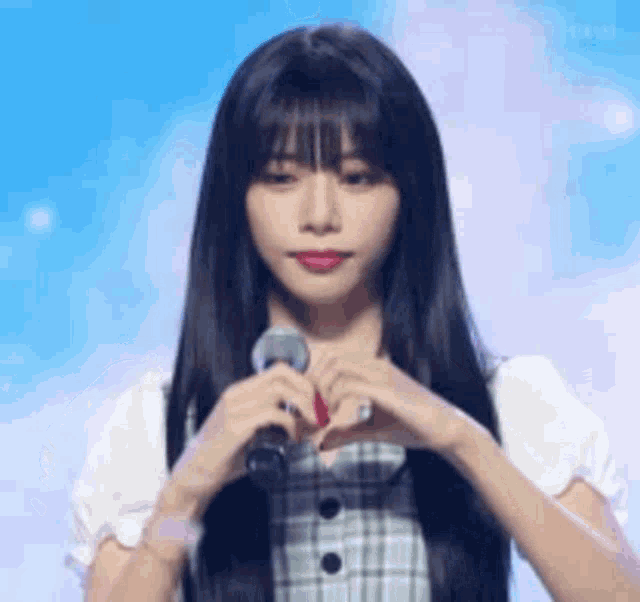 a woman with long black hair is making a heart shape with her hands while standing in front of a microphone .