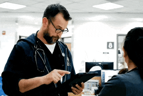 a man with a stethoscope around his neck looks at a tablet while a woman looks on