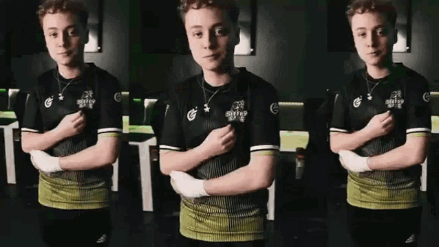 a young man in a black and green shirt is standing with his arms crossed in a dark room .