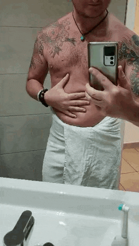 a shirtless man wrapped in a towel is taking a selfie in front of a bathroom mirror .