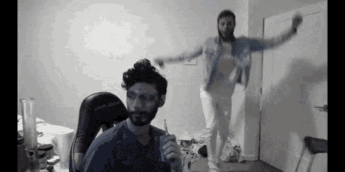 a man with a beard is smoking an electronic cigarette while another man is jumping in the air in a room .