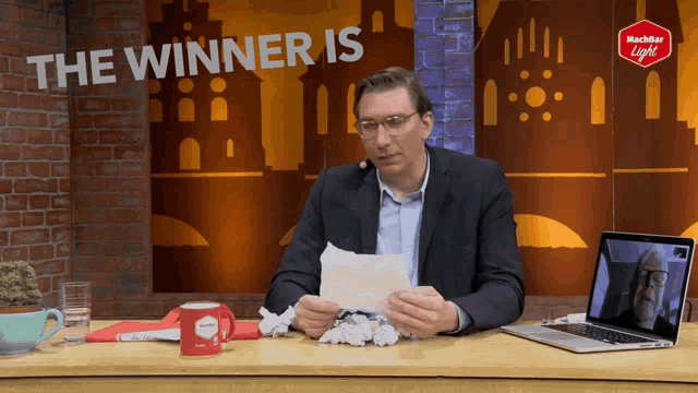 a man is sitting at a desk with a laptop and a sign that says the winner is