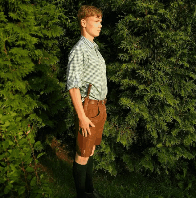 a young man in a green plaid shirt and brown shorts stands in front of a tree