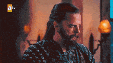 a man with long hair and a beard looks at the camera with a tv logo in the background