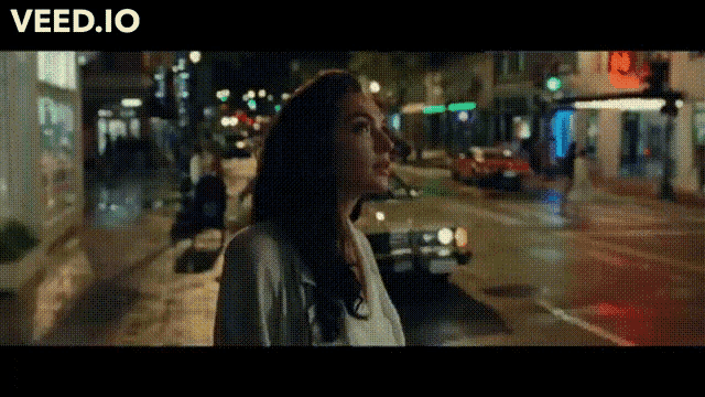 a woman is walking down a city street at night with the website veed.io visible in the corner