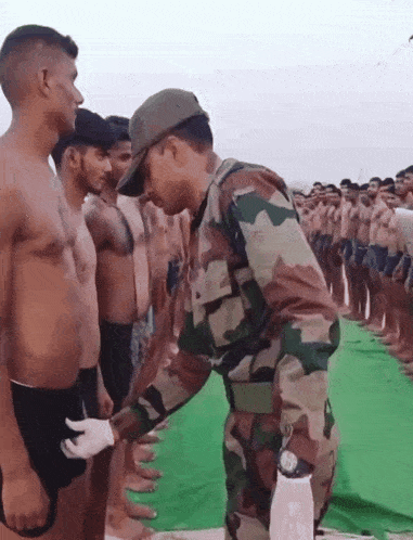 a man in a camouflage uniform stands in front of a group of men without shirts