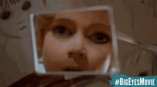 a woman is looking at her face in a mirror with #bigeyesmovie written on the bottom right