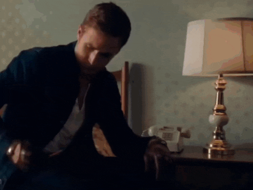 a man is sitting on a bed next to a lamp and a phone .