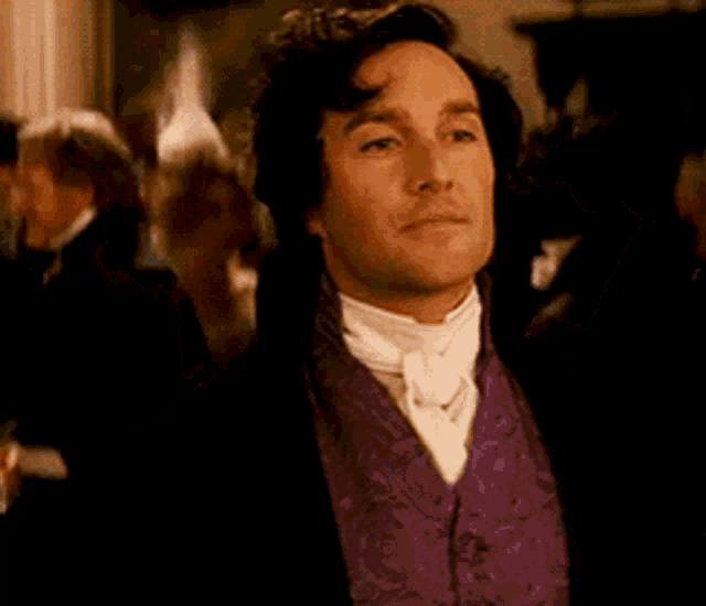 a man in a purple vest and black jacket is standing in front of a crowd of people .