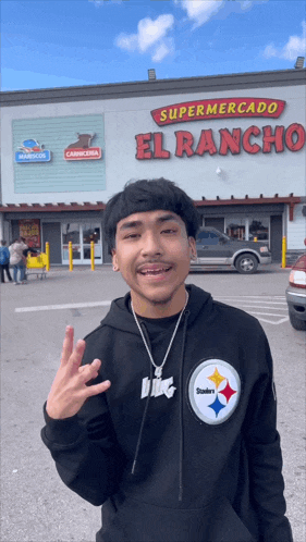 a man wearing a black steelers sweatshirt stands in front of a supermarket called el rancho