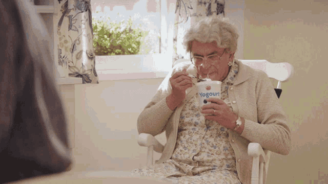 an elderly woman sits in a rocking chair eating yogurt from a cup