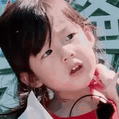 a little girl in a red shirt is looking at the camera with a serious look on her face .