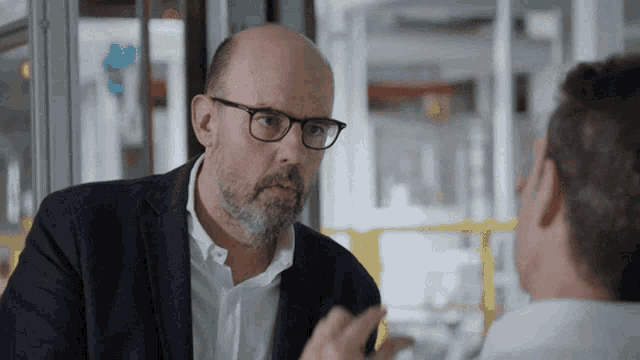 a bald man with glasses talks to another man