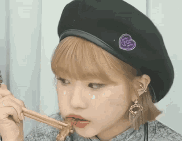 a girl wearing a black beret with a heart on it is eating something with chopsticks