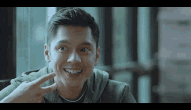 a young man in a green hoodie is smiling and pointing to his face .