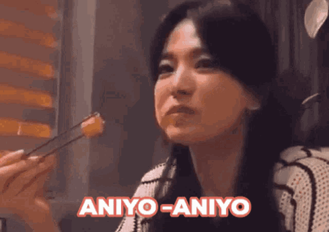 a woman is holding a piece of food with chopsticks and the words aniyo-aniyo are above her