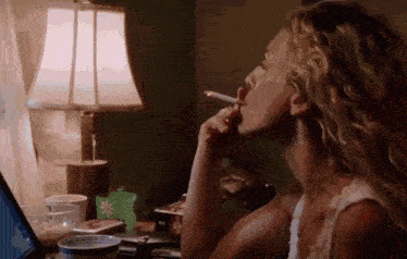 a woman is smoking a cigarette in front of a lamp and a laptop