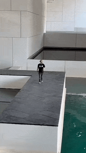 a man wearing a black shirt that says ' a ' on it walks across a ledge overlooking a pool