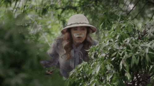 a woman wearing a safari hat and a mask on her face looks through a bush